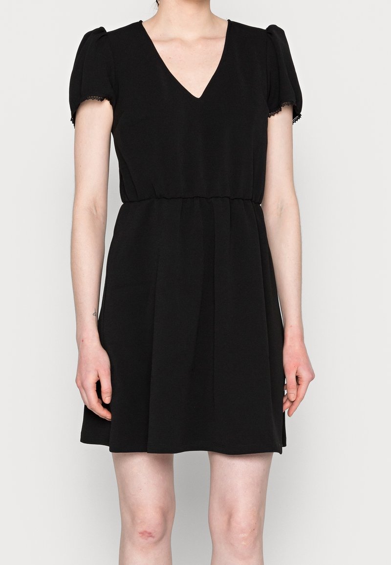 Femme portant une courte robe noire à manches bouffantes et un col en V, debout devant un fond clair uni.