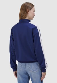 Granatowy sweatshirt z wysokim kołnierzem, białymi paskami po bokach i ściągaczami przy mankietach. Tkanina wydaje się gładka i lekko błyszcząca. Pokazano widok z tyłu.