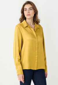 Musztardowa bluzka z długim rękawem, z wywijanym kołnierzykiem, zapinana na guziki, o gładkiej, satynowej fakturze. Dopasowany krój z mankietami.
