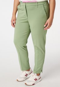 Lichtgroene broek met een getailleerde pasvorm, voorzien van voorplooien en zijzakken. Gecombineerd met witte sneakers met maroon accenten.