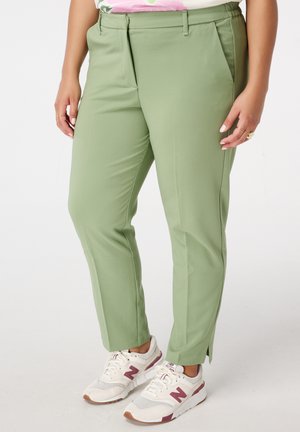 Lichtgroene broek met een getailleerde pasvorm, voorzien van voorplooien en zijzakken. Gecombineerd met witte sneakers met maroon accenten.