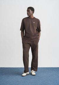 Young man stands on blue carpet wearing brown Adidas tracksuit with white stripes and white sneakers, hands in pockets, looking left.