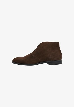 Botines de ante marrón con punta redondeada, que cuentan con tres ojetes para los cordones y una suela de goma negra texturizada.