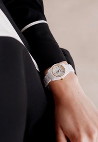Weißes Silikon-Armbanduhr mit einem runden weißen Zifferblatt, goldenen Akzenten und texturiertem Armband. Am Handgelenk getragen, teilweise sichtbar gegen ein dunkles Hemd.