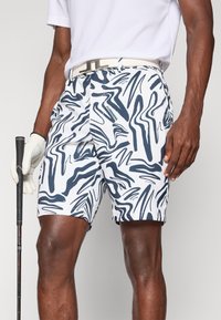 Homem vestindo calções de golfe com padrão abstrato em branco e azul-marinho, cinto branco, luva branca, segurando um taco de golfe com a mão direita repousada na coxa.