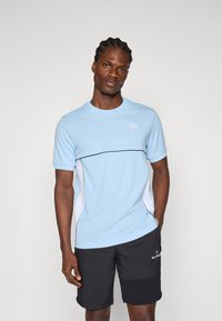 Camiseta deportiva de color azul claro con mangas cortas, ribete negro y acentos laterales blancos; llevada con pantalones cortos negros que presentan un logo.