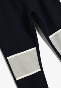 Mörkblå träningsbyxor med grå tygpaneler, med en tunn vit kant. Mjuk textur, smal passform och tapered ben design.