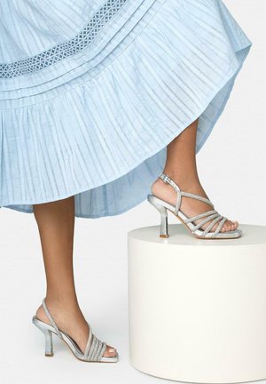 Piernas con sandalias de tacón alto plateadas con tiras, con una falda azul plisada, elevadas sobre un pedestal cilíndrico blanco contra un fondo liso.