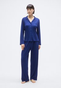 Σετ πιτζάμες σε ναυτικό μπλε χρώμα από απαλό ύφασμα, με μπλούζα με γιακά και κουμπιά μπροστά και μακριά μανίκια, συνδυασμένο με φαρδιά παντελόνια.