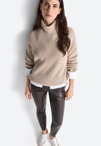 Maglione a coste beige con collo alto indossato sopra una camicia bianca, abbinato a pantaloni in finta pelle marrone e sneakers chiare.