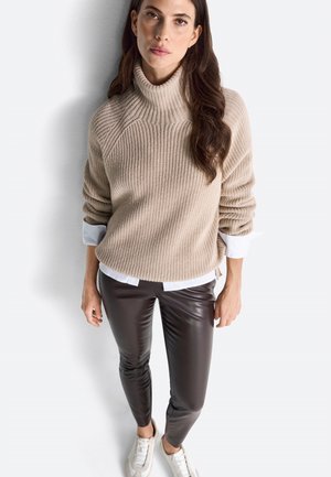 Beige Rippenstrick-Turtleneck-Pullover in Kombination mit einem weißen Hemd, kombiniert mit braunen Kunstlederhosen und hellen Sneakern.