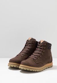 Ruskih nubuk srednje visine, s teksturiranim površinama, čvrstim gumenim đonom i kontrastnom narančastom linijom duž đona. Ima metalne uške i vezice.