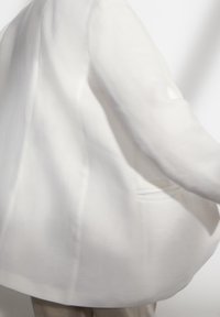 Biały żakiet o miękkiej teksturze, z jedną kieszenią i luźnym krojem. Projekt zawiera lekko opadające ramiona oraz swobodny wygląd.