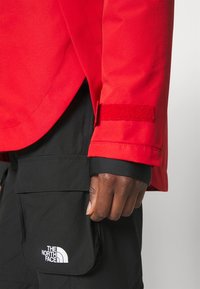 Main foncée levée près de la poche d'un pantalon cargo North Face noir et d'une manche de veste rouge avec sangle Velcro au poignet sur un fond clair.