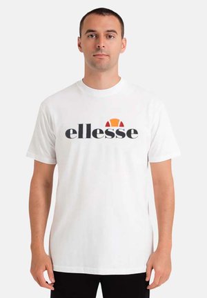 Homme portant un T-shirt blanc avec un logo "ellesse" noir et orange, debout devant un fond uni.