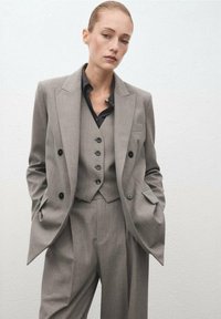 Traje gris a medida con blazer de doble botonadura, chaleco a juego con botones y pantalones de pierna ancha. Tejido texturizado, diseño minimalista.