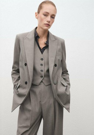 Traje gris a medida con blazer de doble botonadura, chaleco a juego con botones y pantalones de pierna ancha. Tejido texturizado, diseño minimalista.