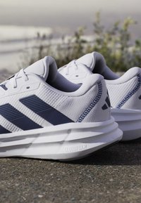 Pair of white and navy running shoes on pavement with textured soles labeled Cloudfoam, blurred plants and sea in the background.