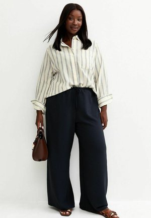 Femme portant une chemise ample à rayures avec boutons, un pantalon large noir, des sandales marron, tenant un sac à main marron foncé sur un fond uni.