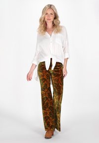 Camicia bianca a bottoni annodata in vita, abbinata a pantaloni in velluto verde e arancione con un vivace motivo paisley. Scarpe casual completano il look.