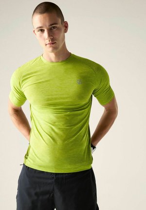 Joven con el pelo corto está de pie con las manos detrás de la espalda, vistiendo una camiseta deportiva de manga corta de color verde brillante y pantalones cortos negros.