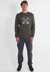 Chemise à manches longues vert olive avec un imprimé graphique comprenant des motifs de feuilles blanches et turquoise ainsi que du texte. Associée à un pantalon gris foncé et des chaussures noires.