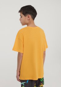 T-shirt di cotone giallo con collo a giro e maniche corte. La vista posteriore rivela una vestibilità rilassata e una texture liscia, abbinata a pantaloni corti a fantasia.