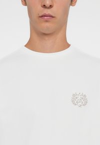 Hvit t-skjorte med rund hals laget av myk bomull, med et detaljert grått brodert logo innenfor et rundt blomsterdesign på brystet.