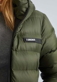 Olijfgroene gewatteerde jas met een matte afwerking, voorzien van een rits en een klein zwart etiket met de tekst "JACKIT" op de borst.