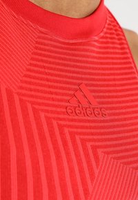 Czerwony sportowy top z wysokim kołnierzem, ozdobiony teksturowanym, paskowanym wzorem oraz wypukłym logo Adidas w dopasowanym kolorze.