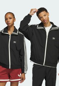Schwarze Windbreaker mit weißen Akzenten, hochgeschlossen, mit Reißverschluss und drei weißen Streifen an den Ärmeln. Modell trägt rote Shorts.