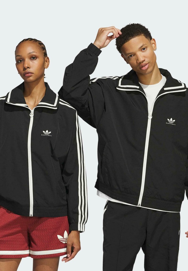 Schwarze Windbreaker mit weißen Akzenten, hochgeschlossen, mit Reißverschluss und drei weißen Streifen an den Ärmeln. Modell trägt rote Shorts.