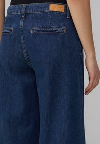 Pantalons en denim bleu marine à jambe large avec taille haute. Comprend deux poches arrière et une patch en cuir sur la ceinture. Texture lisse.