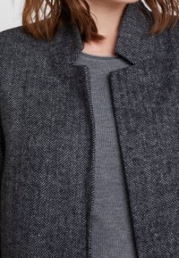 Gros plan sur une personne portant un manteau gris foncé à chevrons par-dessus un pull côtelé gris clair.