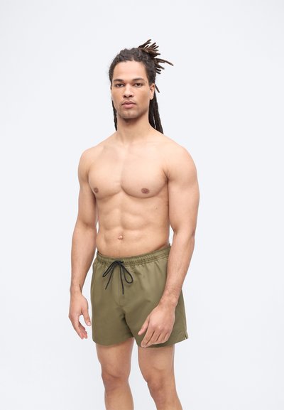 Άνδρας χωρίς πουκάμισο με μακριές πλεξούδες να στέκεται, φορώντας χακί πράσινο μαγιό με μαύρο κορδόνι, μπροστά σε ένα απλό λευκό φόντο.