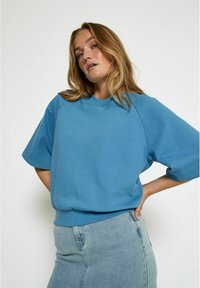 Felpa oversized blu con scollo rotondo e maniche corte, abbinata a una gonna di jeans blu chiaro. Tessuto morbido e liscio.