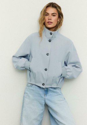 Młoda kobieta w jasnoniebieskiej, zapinanej na guziki kurtce z wysokim kołnierzem oraz jasnoniebieskich dżinsach, stojąca na tle jednolitego, białego tła.