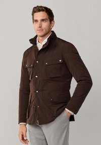 Mand iført en mørkebrun jakke med fire knap-lukket lommer, hvid skjorte og beige bukser, stående med den ene hånd i lommen.