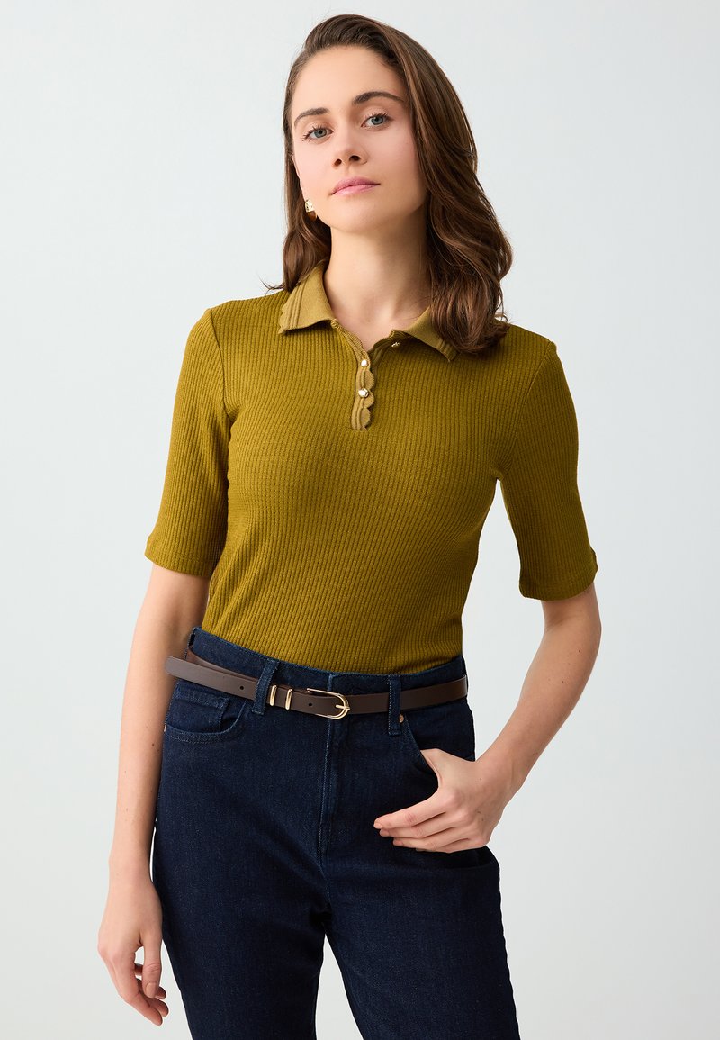 Jeune femme aux cheveux bruns portant un polo côtelé couleur moutarde à manches courtes et un jean taille haute bleu foncé avec une ceinture marron.