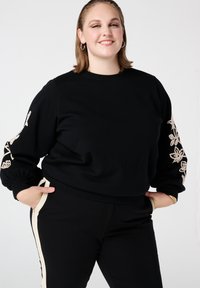 Zwart cropped sweatshirt met geborduurde bloemaccenten op de mouwen. Het materiaal is zacht en heeft een ontspannen pasvorm. Draagt zwarte broek met zijstrepen.