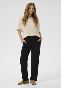 Top scurt cu mânecă scurtă, texturat, de un bej deschis, asociat cu pantaloni negri largi. Pantofi slip-on de nuanță bej, stând cu mâinile în buzunare, fundal neutru.