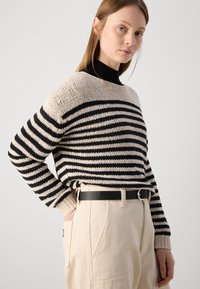 Striped genser i beige og svart, tykk strikk, vide ermer, rund hals; kombinert med lyse beige bukser og et svart belte.