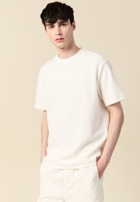 Vit kortärmad t-shirt i ett mjukt, strukturerat tyg med rund halsringning och subtila sidodetaljer. Bärs med ljusa byxor.