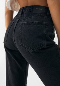 Jeans de denim negro con cintura alta, tejido texturizado y una etiqueta con logo en la parte trasera. Incluye dos bolsillos traseros con detalles de costura.