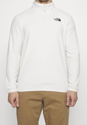 Mężczyzna ubrany w biały polar na ćwierć zamka z logo The North Face oraz beżowe spodnie typu cargo, stojący na jednolitym tle.