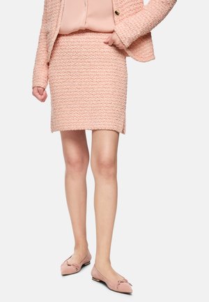 Frau trägt ein rosa strukturiertes Kostüm mit passender Bluse und rosa spitzen Ballerinas mit Schleifen, steht vor einfarbigem Hintergrund.