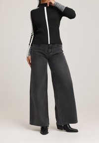 Femme portant un jean noir à jambes larges, un pull à manches longues noir et blanc, et des bottines noires à bout pointu, posant avec une main près du visage.