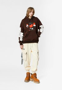 Joven con sudadera marrón con estampado gráfico, pantalones cargo crema con bolsillos grandes y botas de cordones color camel, de pie sobre un fondo blanco.