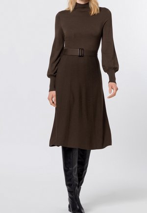 Mujer que lleva un vestido midi marrón de manga larga con cinturón, combinado con botas de cuero negras hasta la rodilla, sobre un fondo liso.