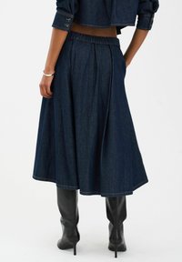 Tamna traper suknja s elastičnom kombinacijom oko struka i nabranim dizajnom, koja doseže do sredine lista. Uparena s elegantnim crnim čizmama do koljena.